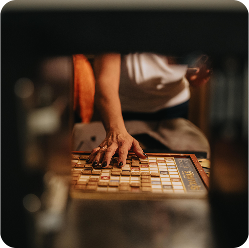 A hand with dark nail polish reaches for a vintage board game, partially visible through an object in the foreground.