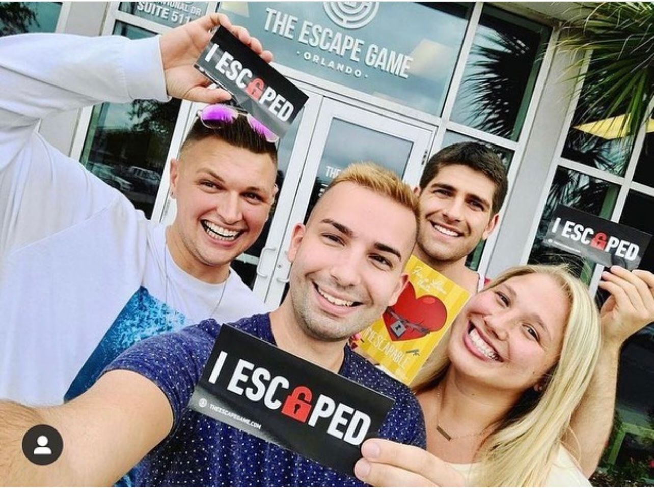 Four smiling friends hold signs reading "I ESCAPED" outside The Escape Game location. The background features the venue's entrance.