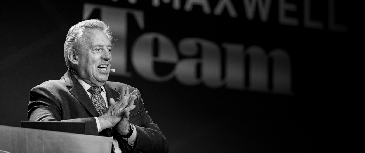 A smiling man in a suit gestures with his hands while speaking at a podium, with a large sign reading "Team" in the background.