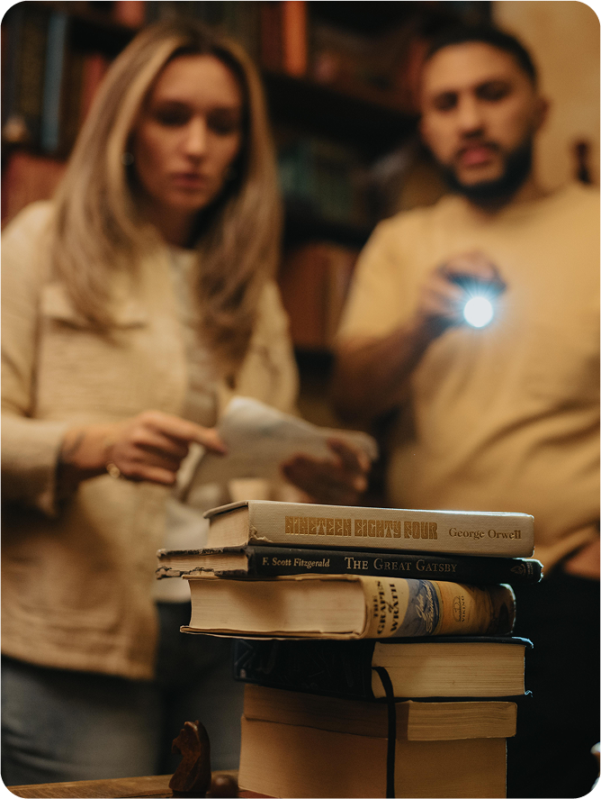 Two people are examining papers, with one holding a flashlight. A stack of books, including "1984" and "The Great Gatsby," is nearby.
