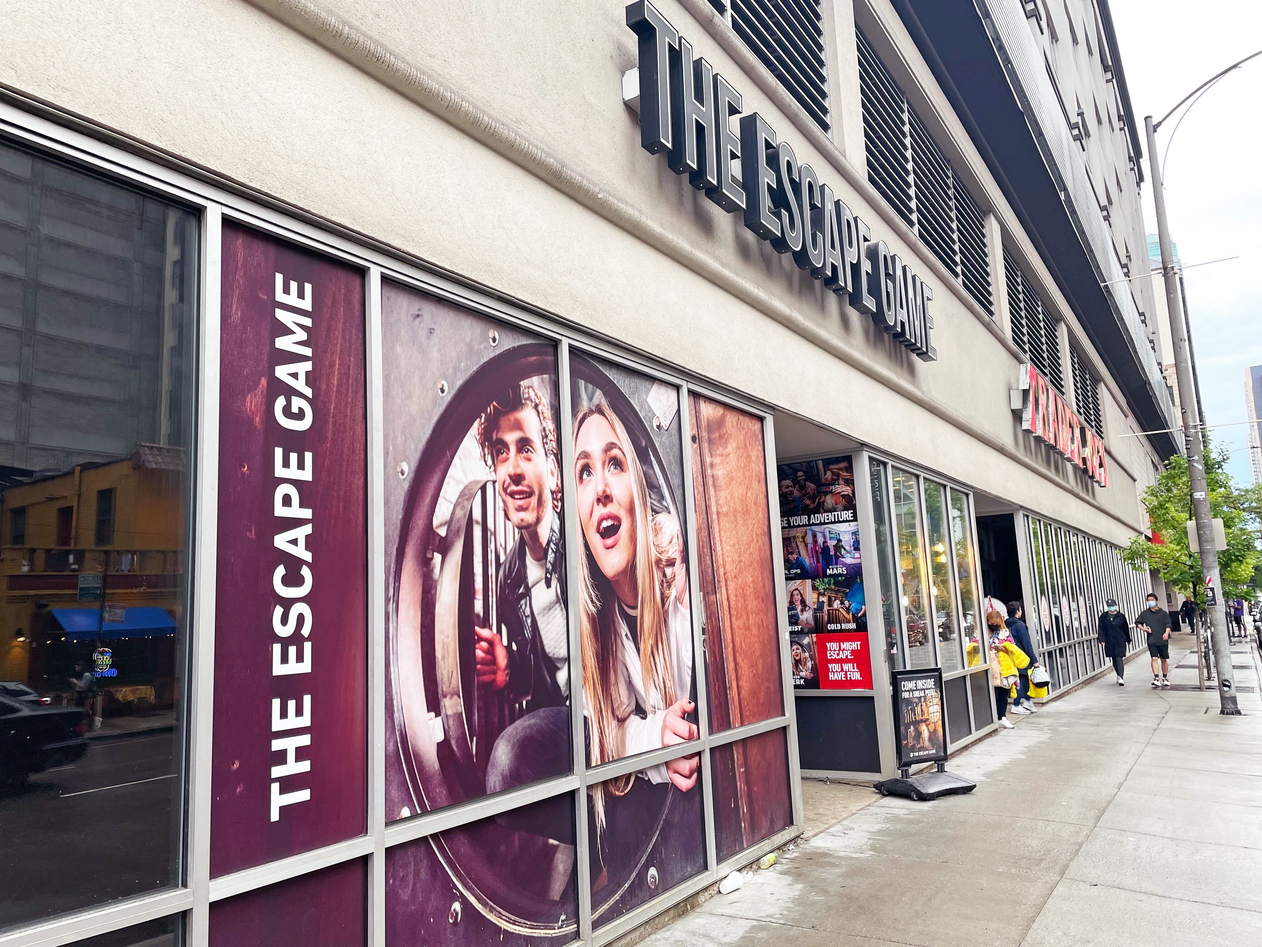 Exterior of "The Escape Game" with large windows displaying excited faces and promotional signs; pedestrians nearby.