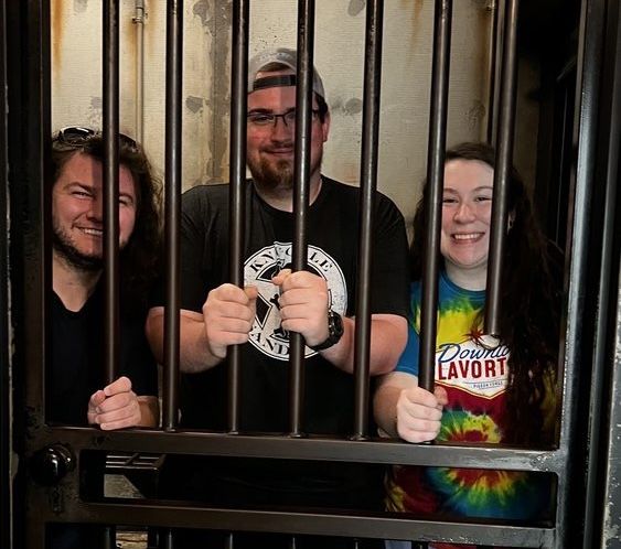 Three young adults smile and pose behind prison bars, appearing playful in a dimly lit environment.