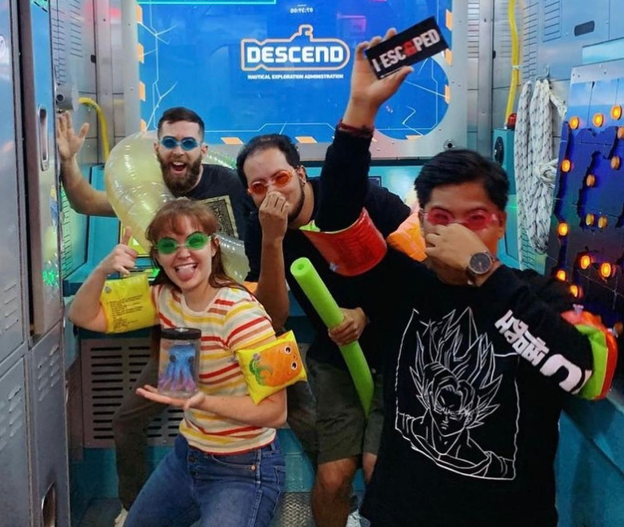 Four people inside a colorful room, wearing swim goggles and inflatable accessories, posing playfully with props.