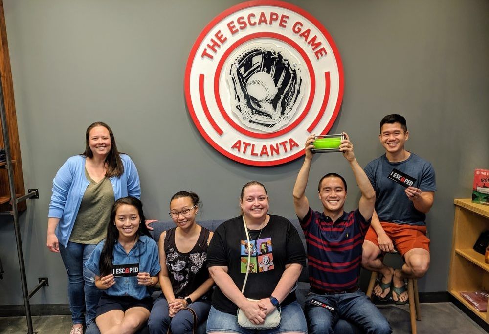 A group of six people smiles in front of a round logo that reads "The Escape Game Atlanta." They celebrate a successful escape.