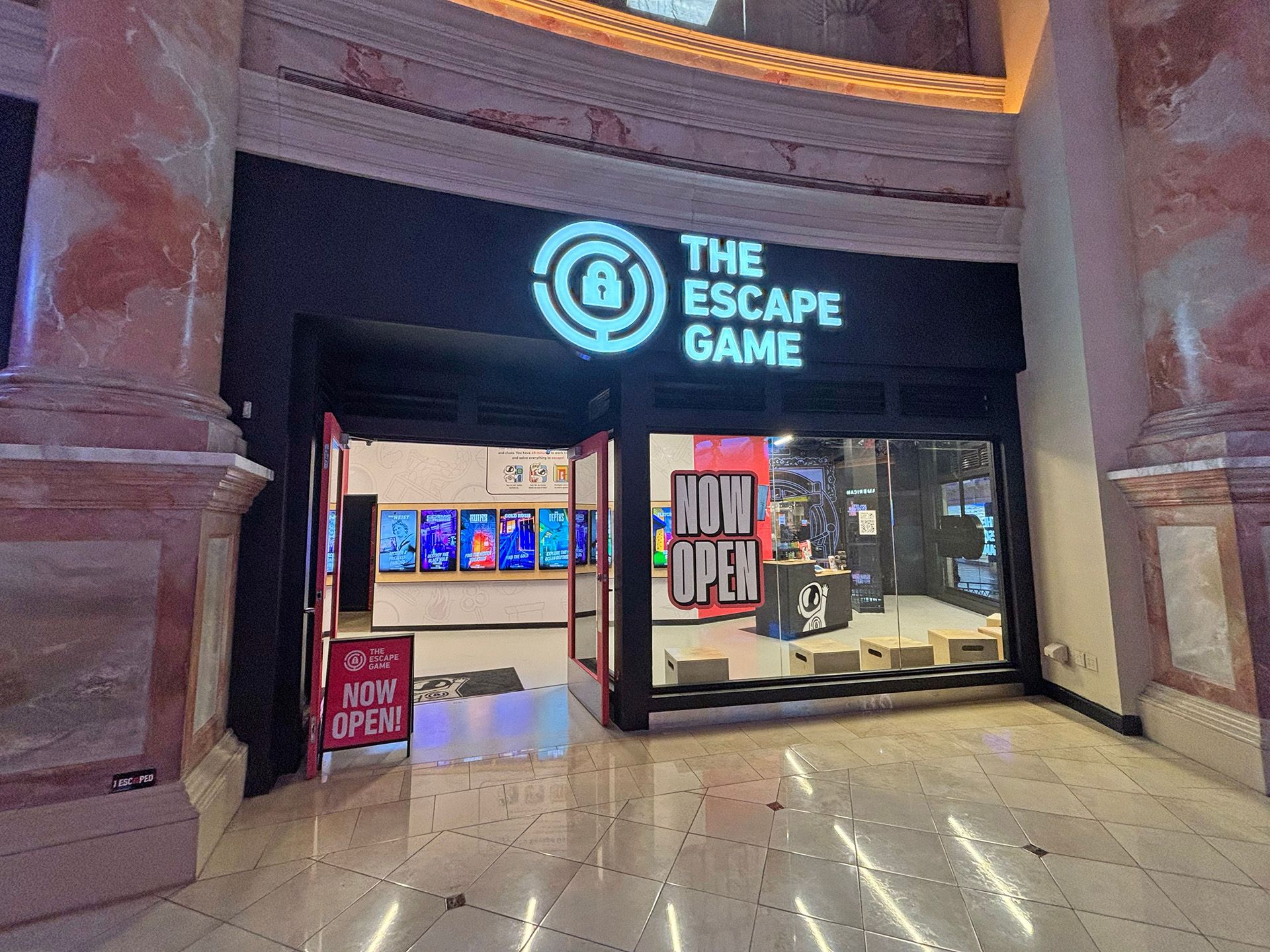 Entrance to "The Escape Game," featuring a large sign, glass doors, and a "NOW OPEN" sign. Brightly lit interior visible.