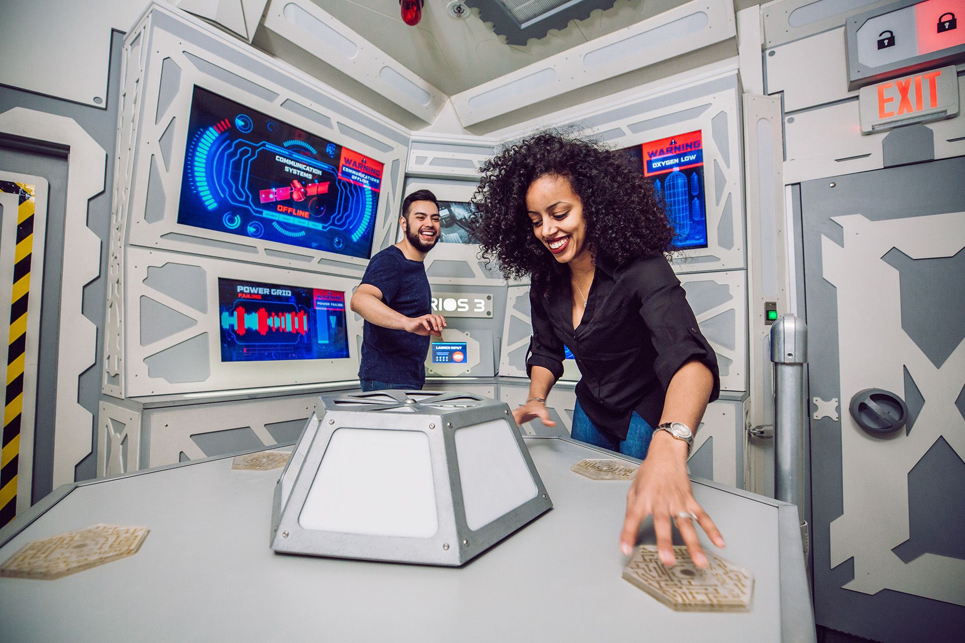 Two people are interacting in a high-tech room with screens displaying digital interfaces. The woman is smiling while touching a central console, and the man is positioned behind her, also engaged.