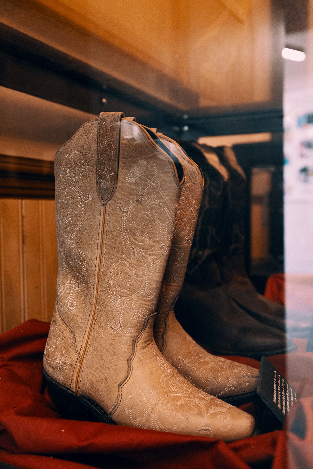 White cowboy boots with intricate embroidery displayed in a glass case, resting on a red fabric backdrop.