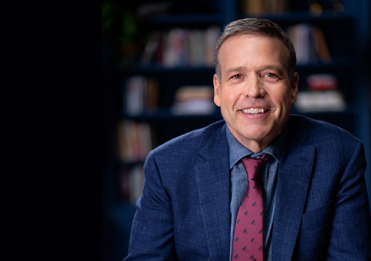 Smiling man in a blue suit and red tie, sitting in front of a blurred bookshelf.