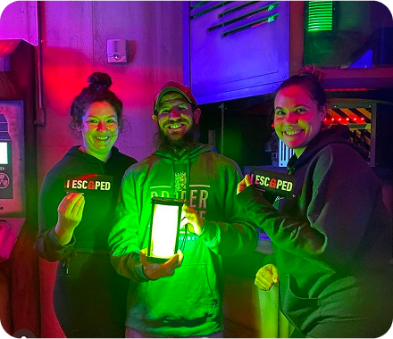 Three people smile in a colorful escape room, holding a "I ESCAPED" sign and a glowing lantern.