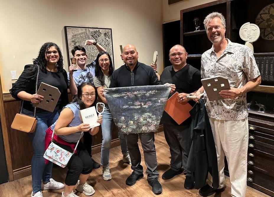 A group of seven people poses together, holding various props like a painting, a book, and game pieces, in a cozy room.