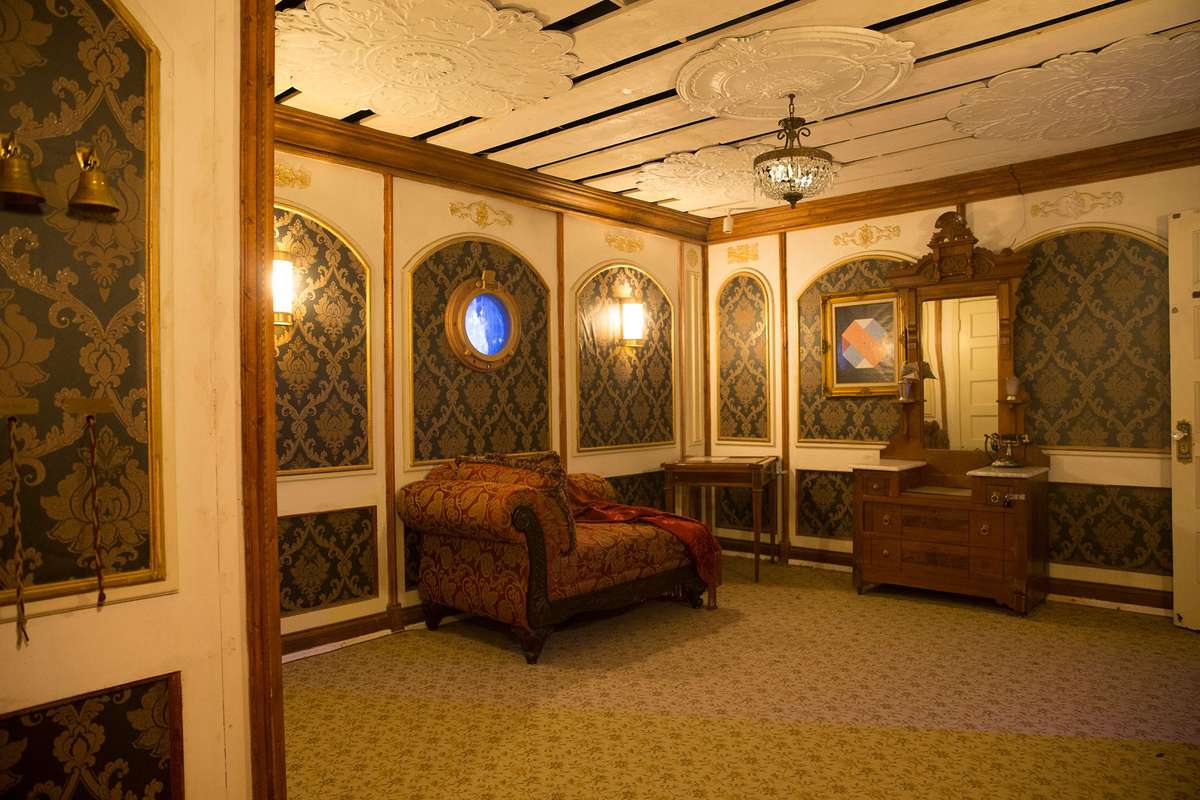 Luxurious room with ornate wallpaper, a plush couch, a wooden mirror, and a round window. Warm lighting enhances the decor.