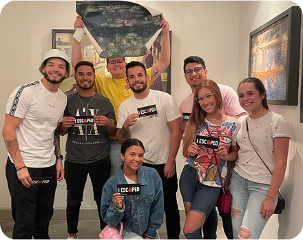 A group of eight people smiling, holding signs that read "I ESCAPED," in a vibrant room with art on the walls.