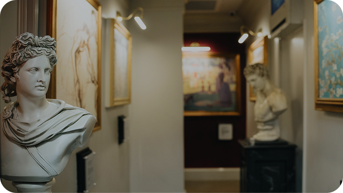 Two busts of classical sculptures stand in a hallway adorned with framed artworks and soft lighting.