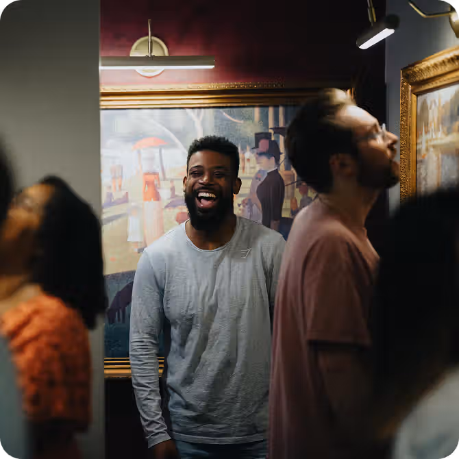 A smiling man in a gray shirt stands among people looking at artwork in a gallery, surrounded by colorful paintings.