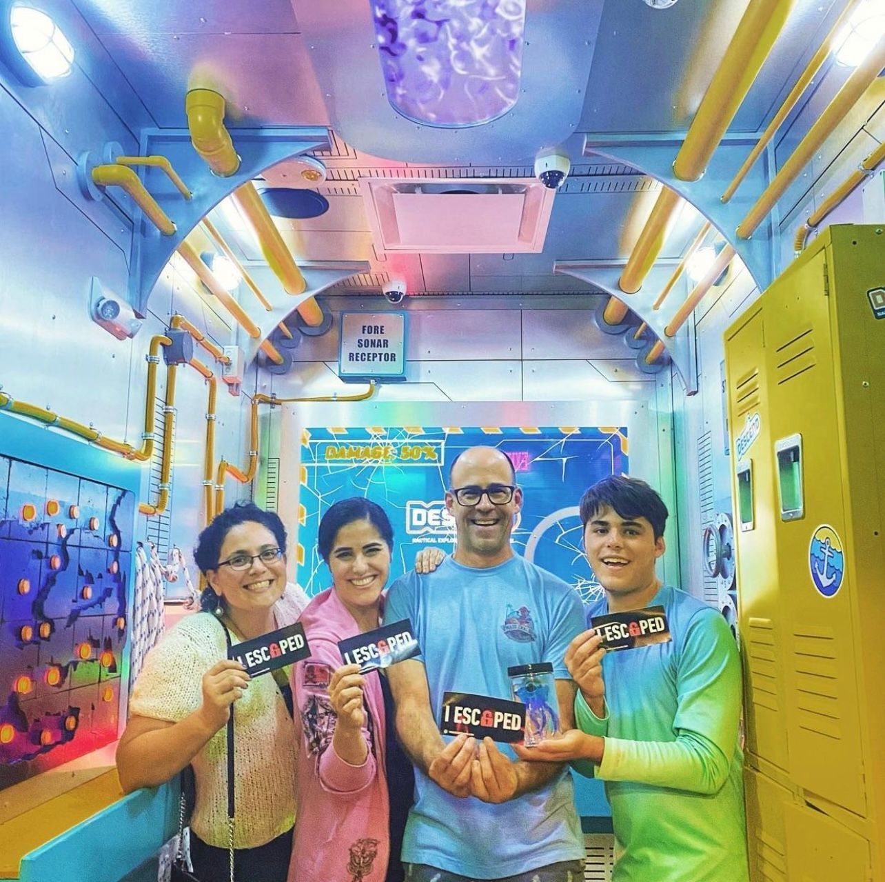 Four people stand in a colorful escape room, each holding a sign that reads "I ESCAPED." Bright, playful decor surrounds them.
