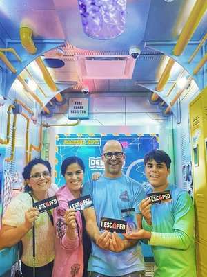 Four people stand in a colorful escape room, each holding a sign that reads "I ESCAPED." Bright, playful decor surrounds them.