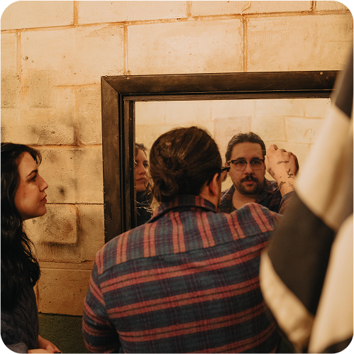 A man with glasses and a plaid shirt writes in a mirror while a woman observes him, both in a rustic setting.