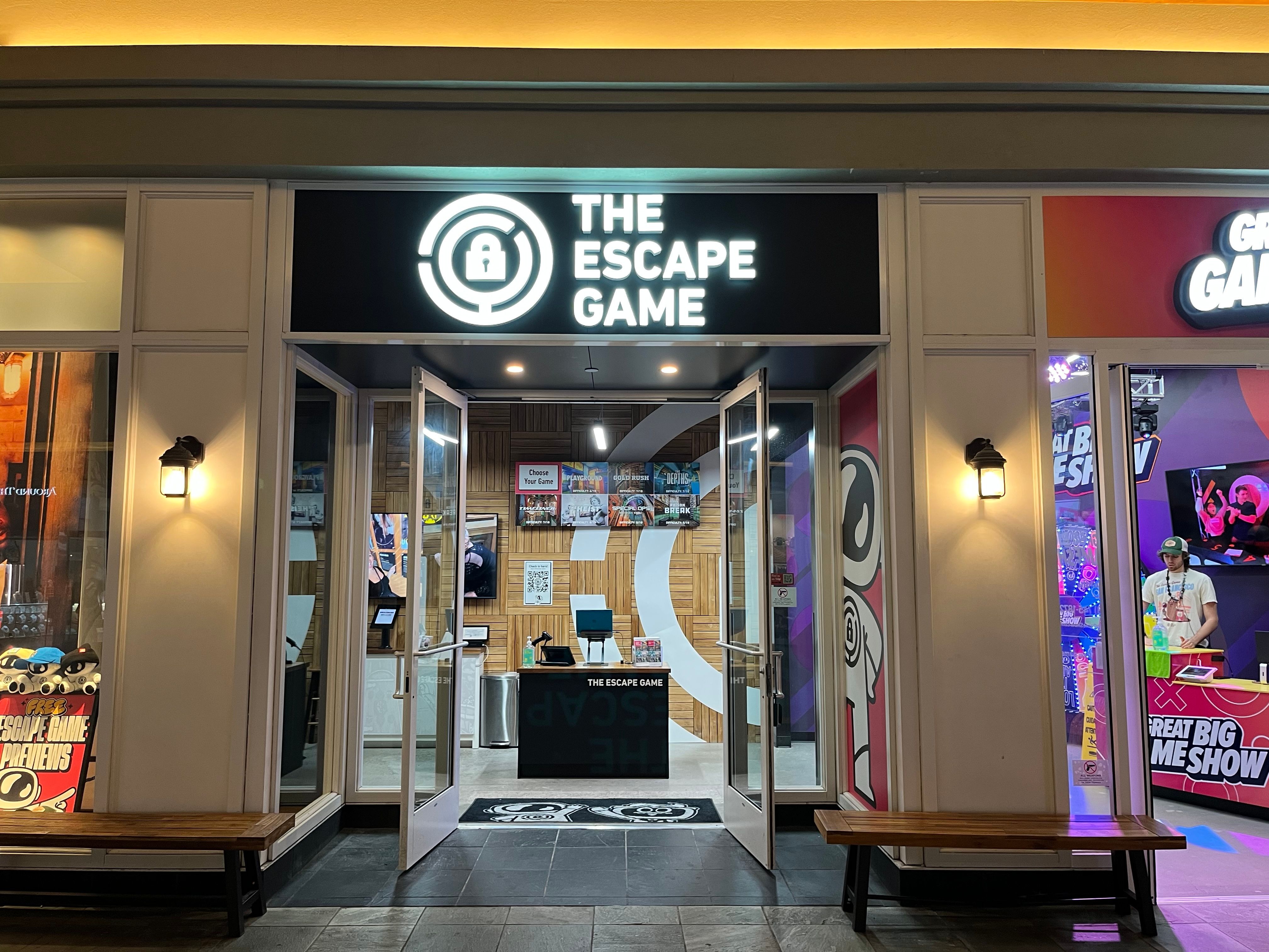 Entrance of "The Escape Game" with a sign above the door and colorful displays inside. Benches are positioned outside.