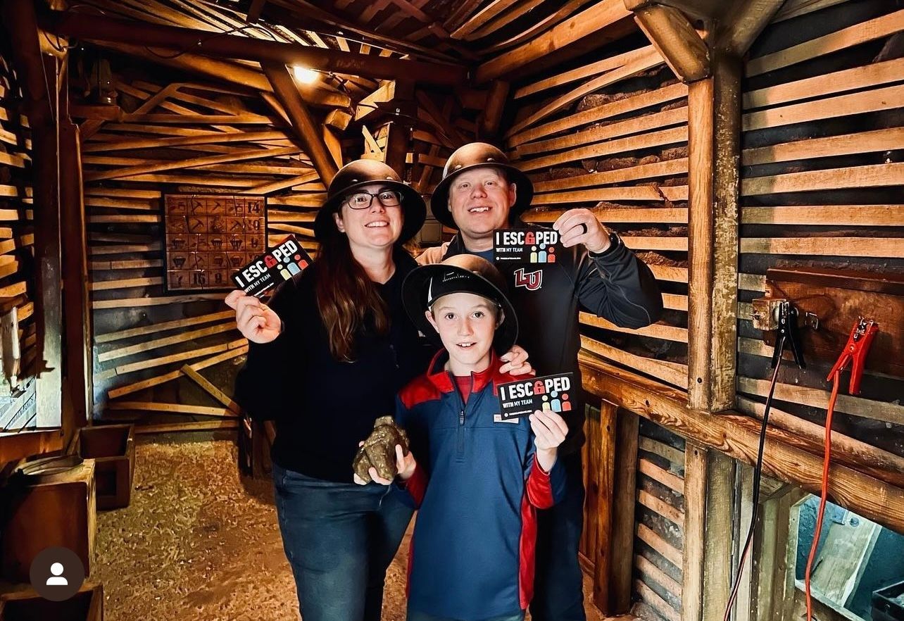 A family of three smiles in a wooden room, each holding an "I Escaped" card. The woman and man wear helmets, and there's a rustic background.