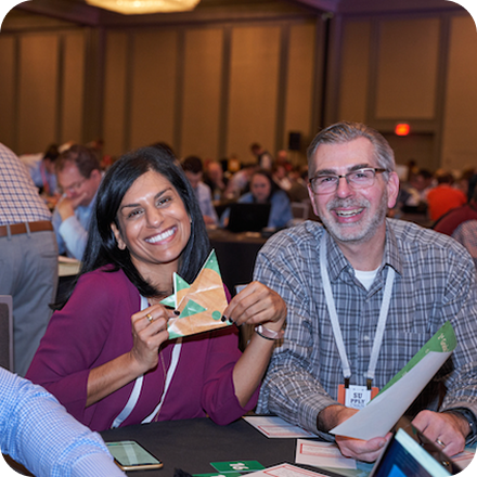 Two smiling individuals are seated at a table in a conference setting. A woman holds an origami piece, while a man next to her smiles broadly.