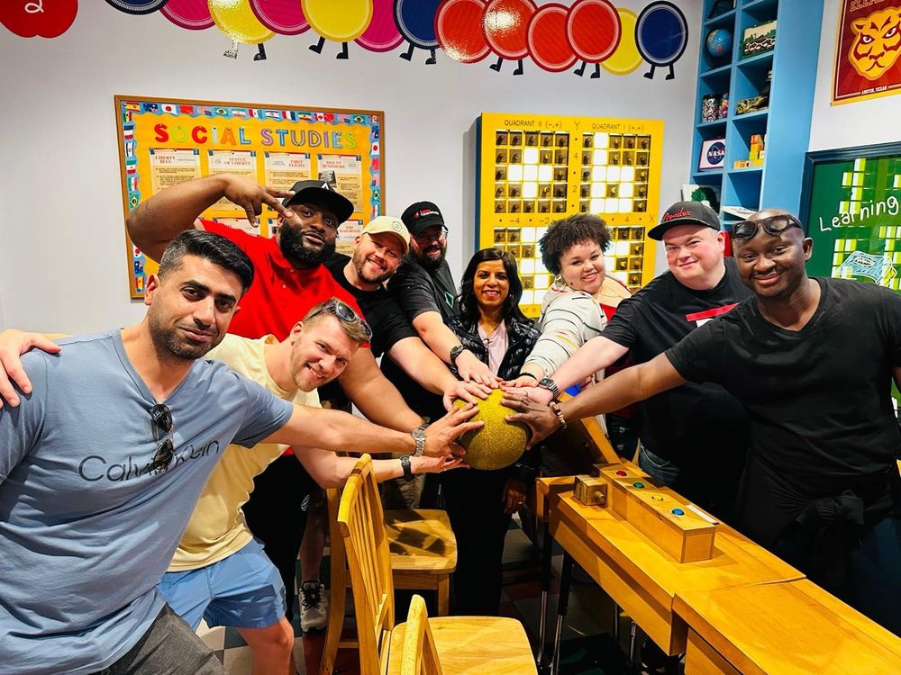 A diverse group of eight people gather around a golden ball, smiling and posing in a colorful classroom setting.