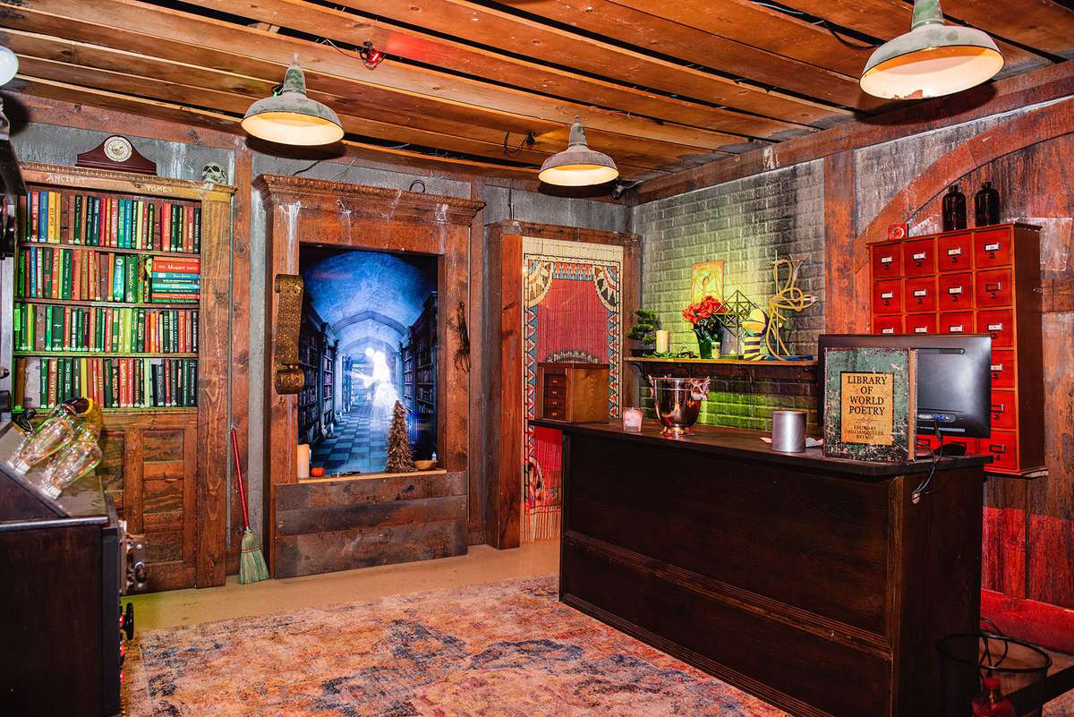 Cozy room with wooden accents, bookshelves, a desk, and a digital display of a library hallway. Warm lighting and decorative items.