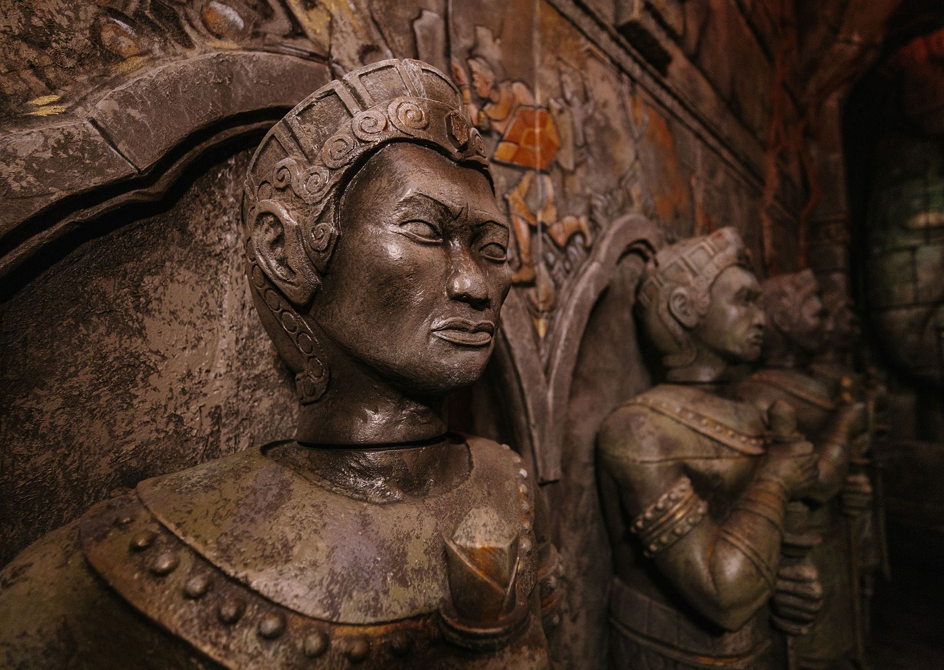Close-up of a detailed stone statue of a warrior with a fierce expression, surrounded by other similar statues in a dimly lit setting.