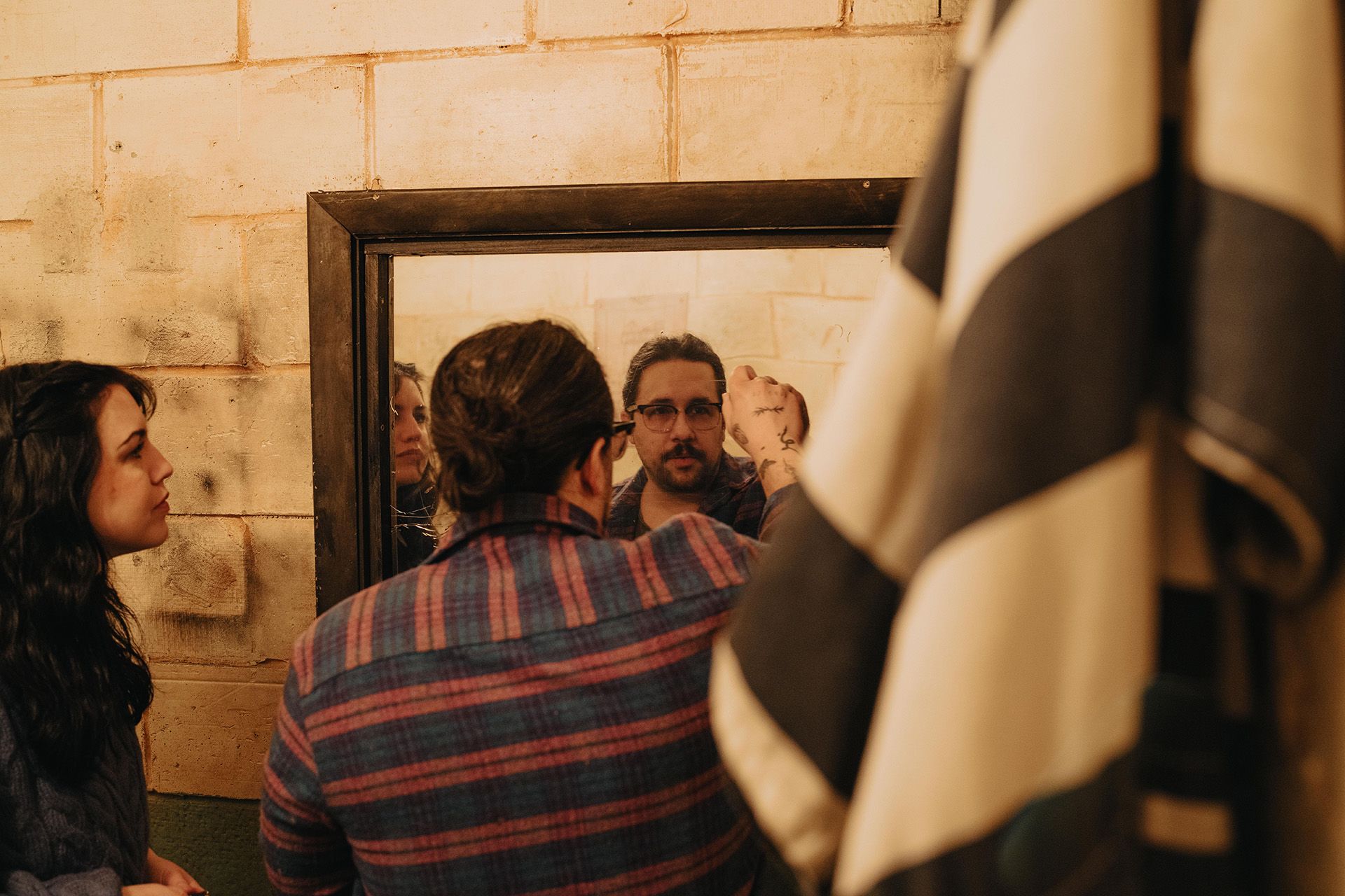 A man with glasses looks into a mirror, while a woman observes him. A striped cloth is partially visible in the foreground.