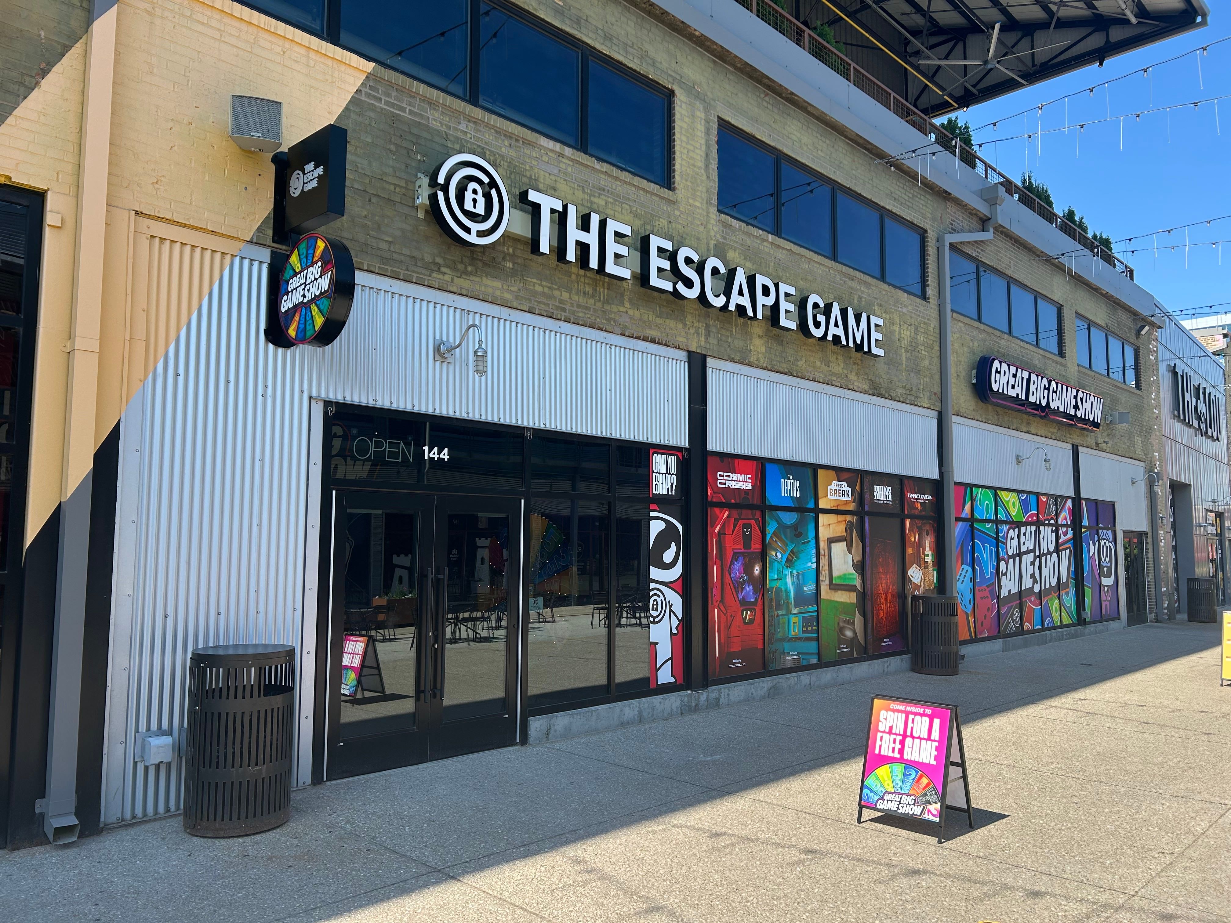Exterior of "The Escape Game" building with colorful graphics, large windows, and an open sign. A sign invites visitors to spin for a free game.