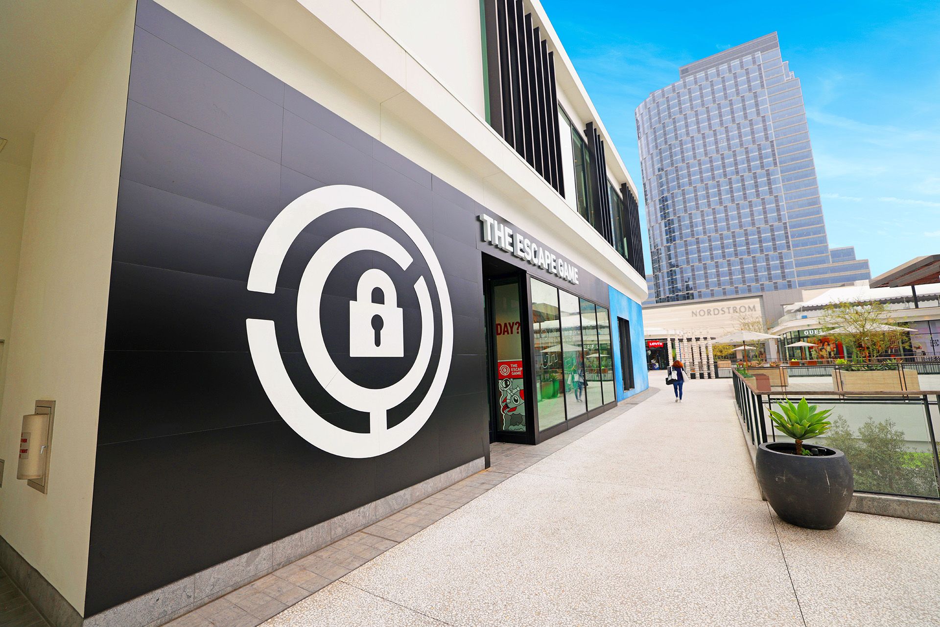 Exterior view of "The Escape Game" storefront, featuring a large lock logo, with a modern building and a shopping area in the background.