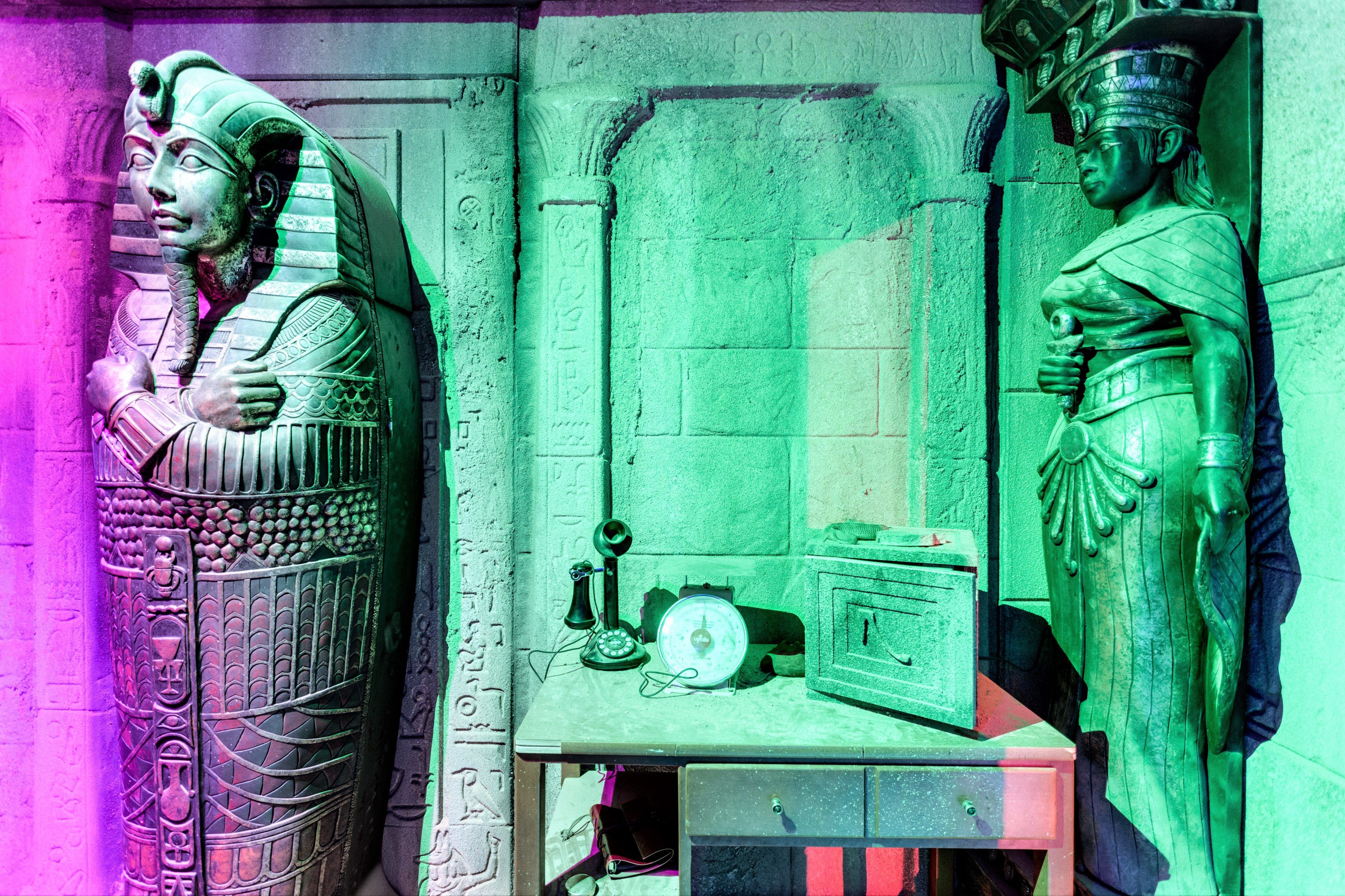 Two ancient Egyptian statues stand beside a table with a clock and a small box, illuminated by colorful lighting.