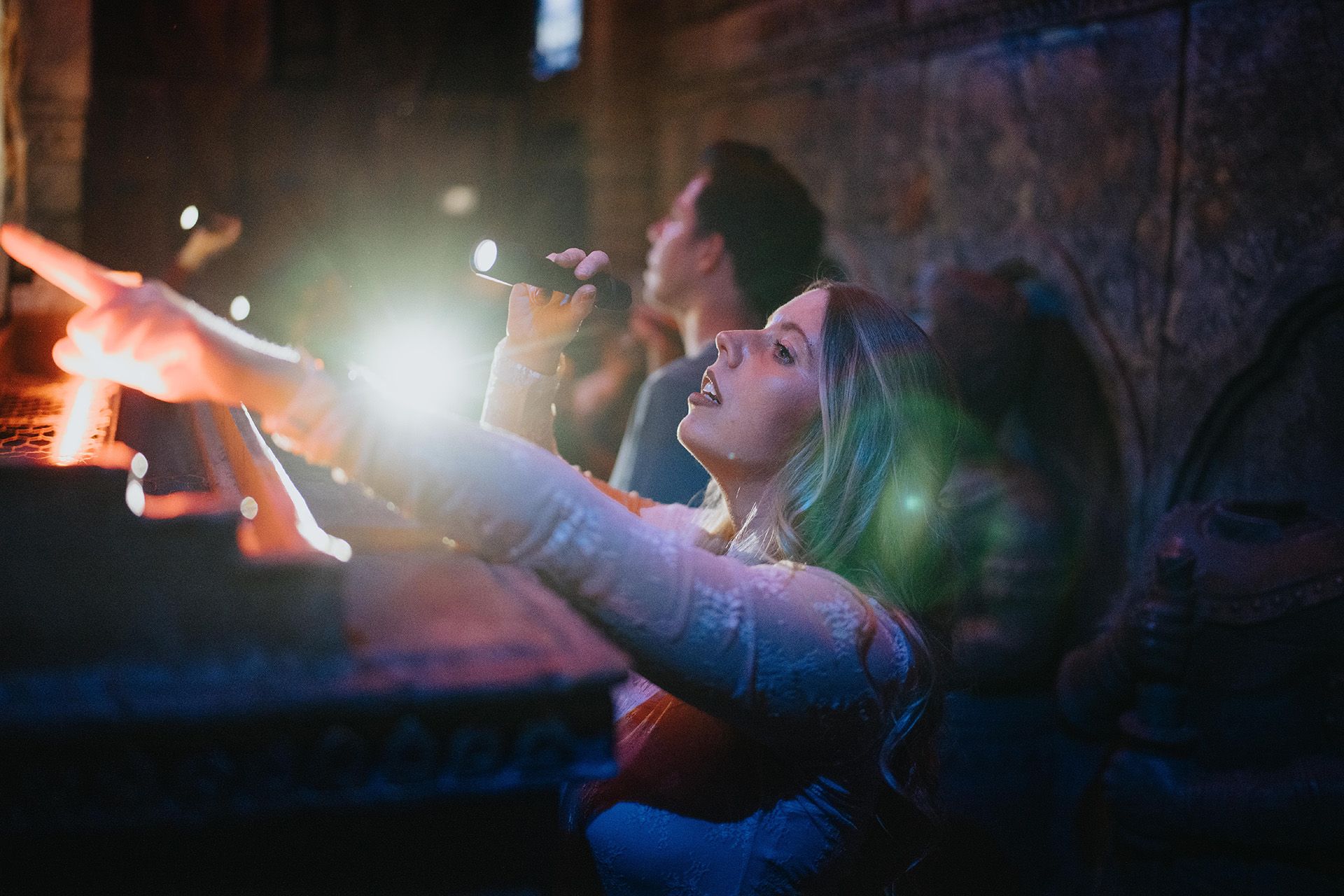 A woman holds a microphone, gesturing expressively while another person stands nearby, both illuminated by flashlight beams.