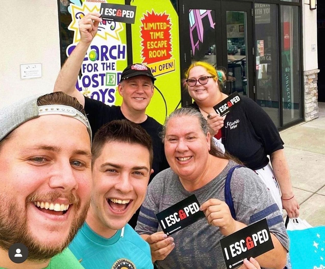 Group of five people smiling in front of an escape room sign, each holding "I ESCAPED" cards. Colorful background.