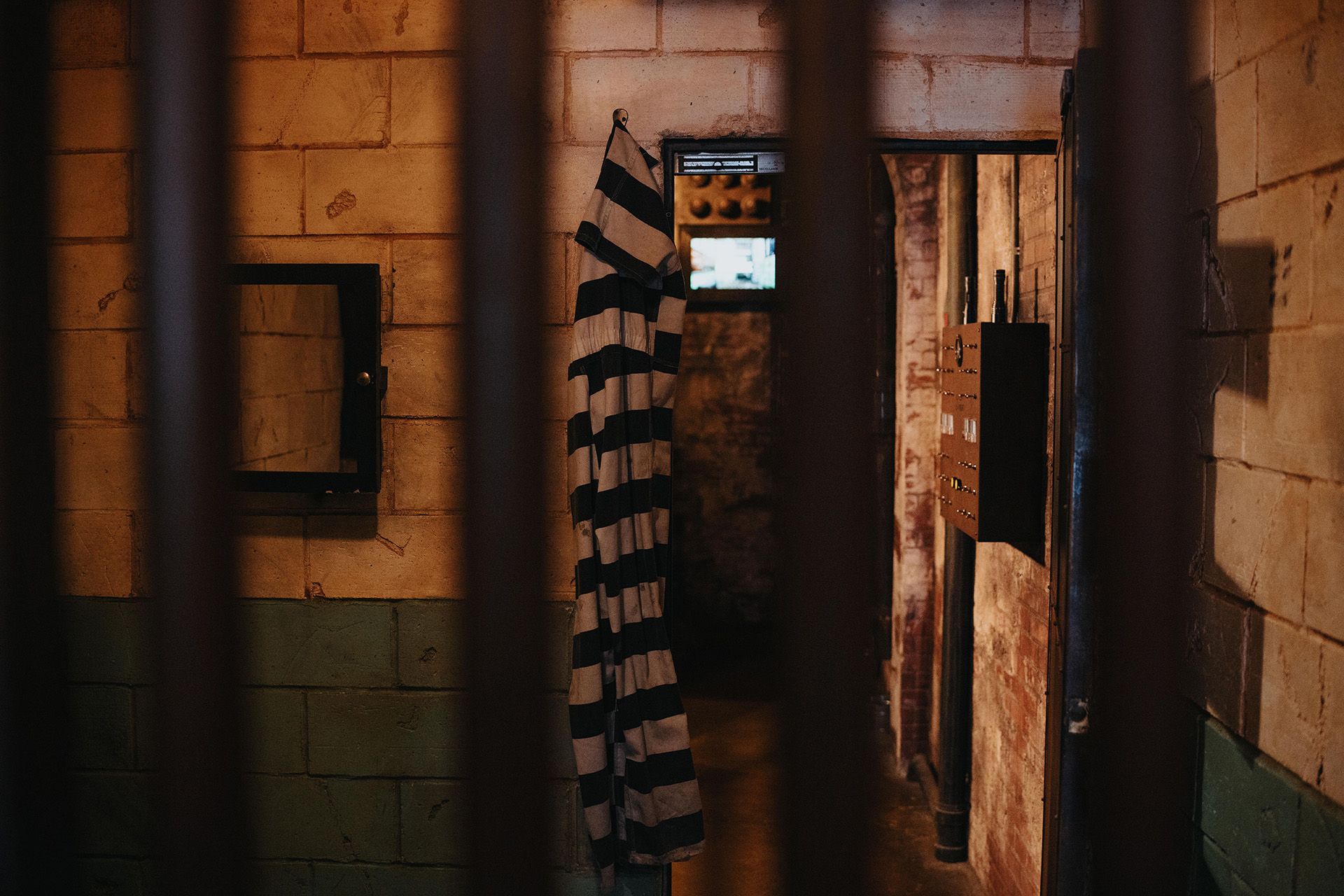 Interior of a prison cell, featuring a barred window, a striped inmate's uniform hanging, and a small mirror on the wall.