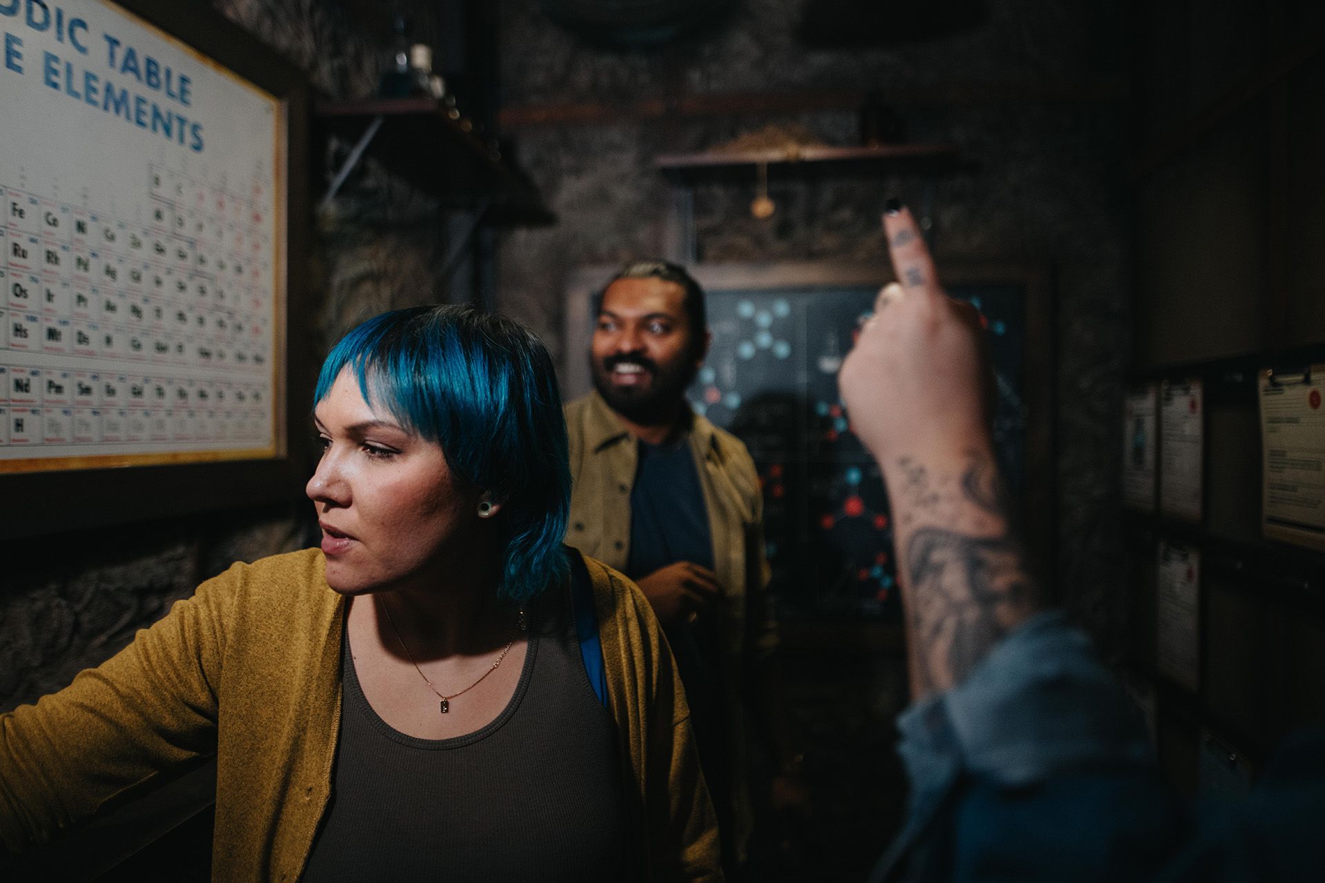 Three people are in a dimly lit room, examining a periodic table of elements on the wall. One woman with blue hair looks closely, while a man smiles in the background. A tattooed hand gestures toward the table.