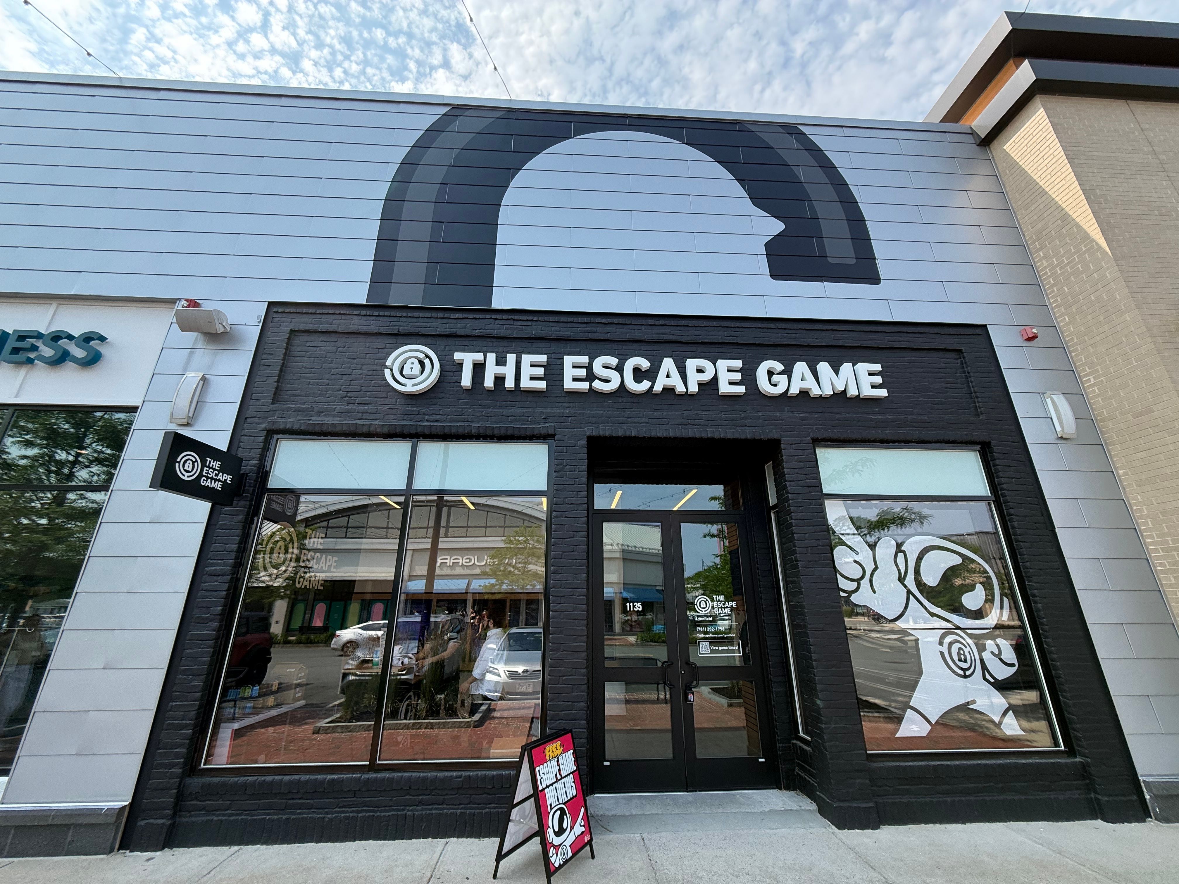 Front of "The Escape Game" store featuring large windows, black signage, and a playful character graphic.