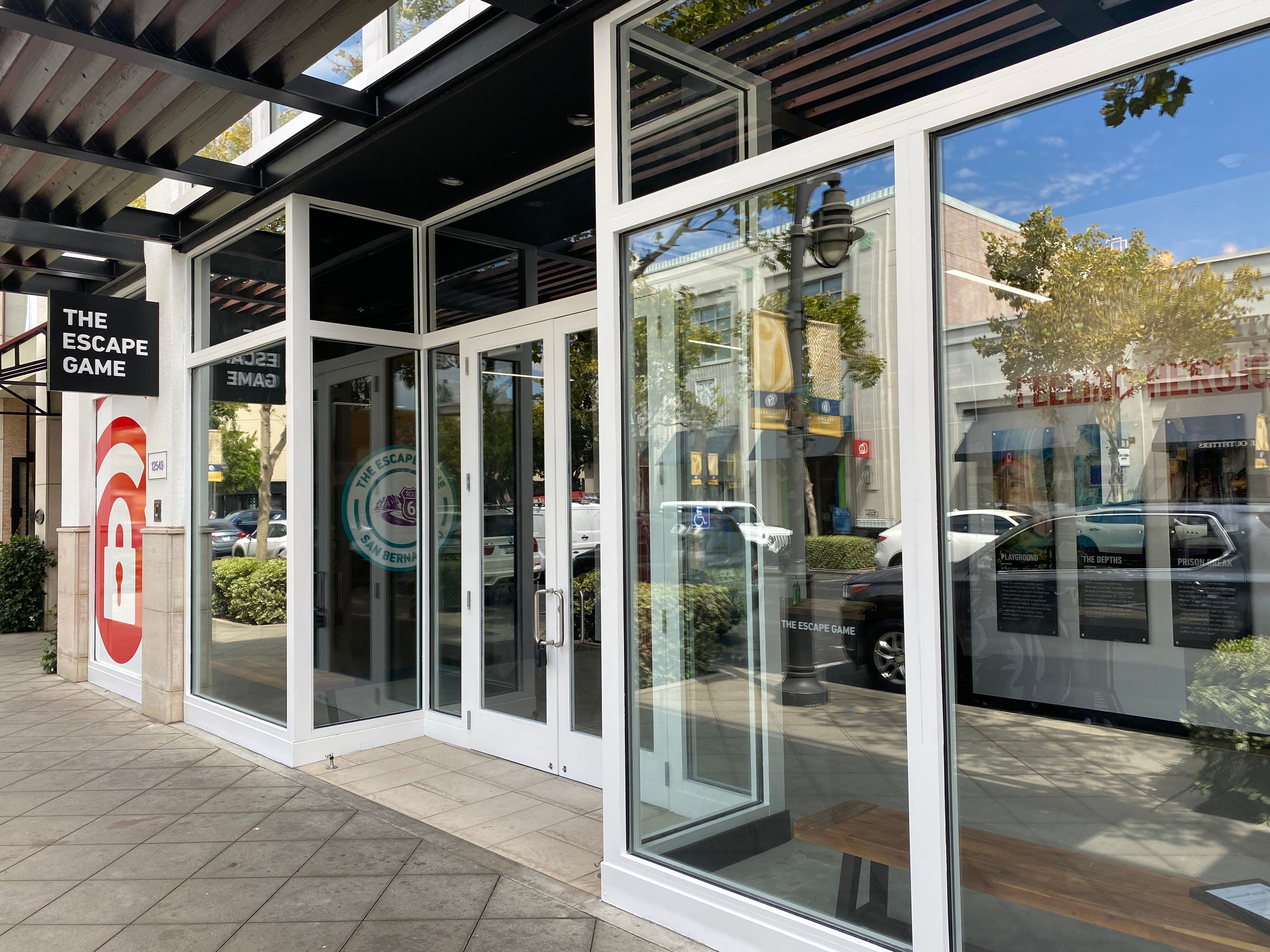 Entrance to "The Escape Game" featuring large glass doors and a sign above. The area is lined with trees and outdoor seating.