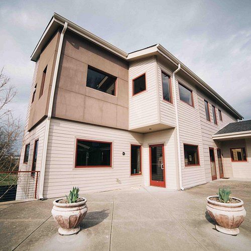 Modern two-story house with a mix of siding and stucco, featuring large windows and potted plants at the entrance.