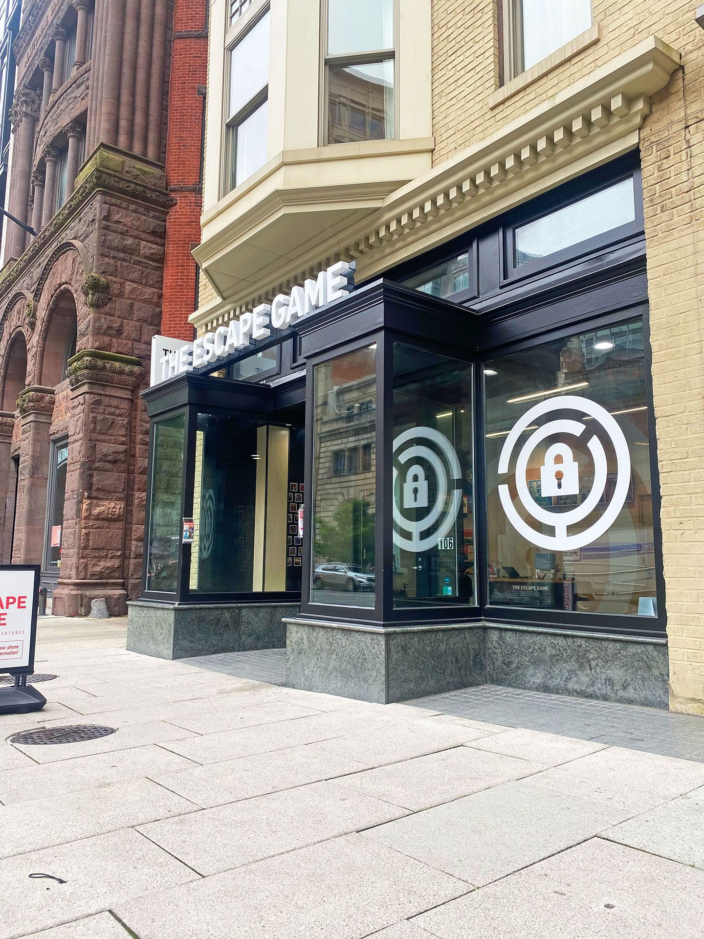 Exterior view of "The Escape Game" storefront, features large windows with lock graphics and a sidewalk sign.