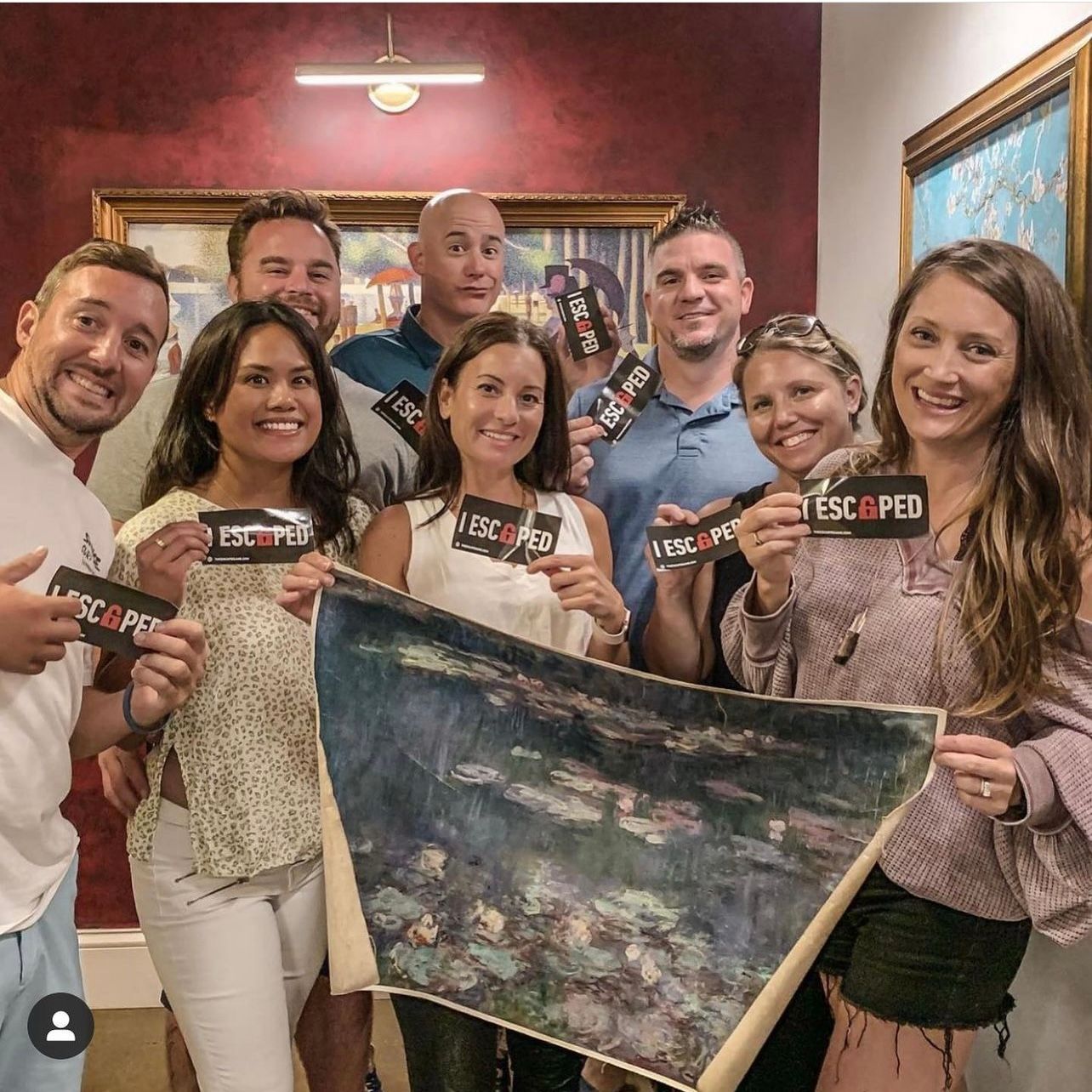 A group of nine people poses together, holding "ESCAPED" stickers and a painting of water lilies. A colorful backdrop adds depth.