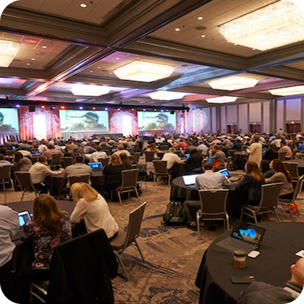 Conference attendees seated in a large hall, focused on a presentation displayed on screens, with colorful lighting and tables.