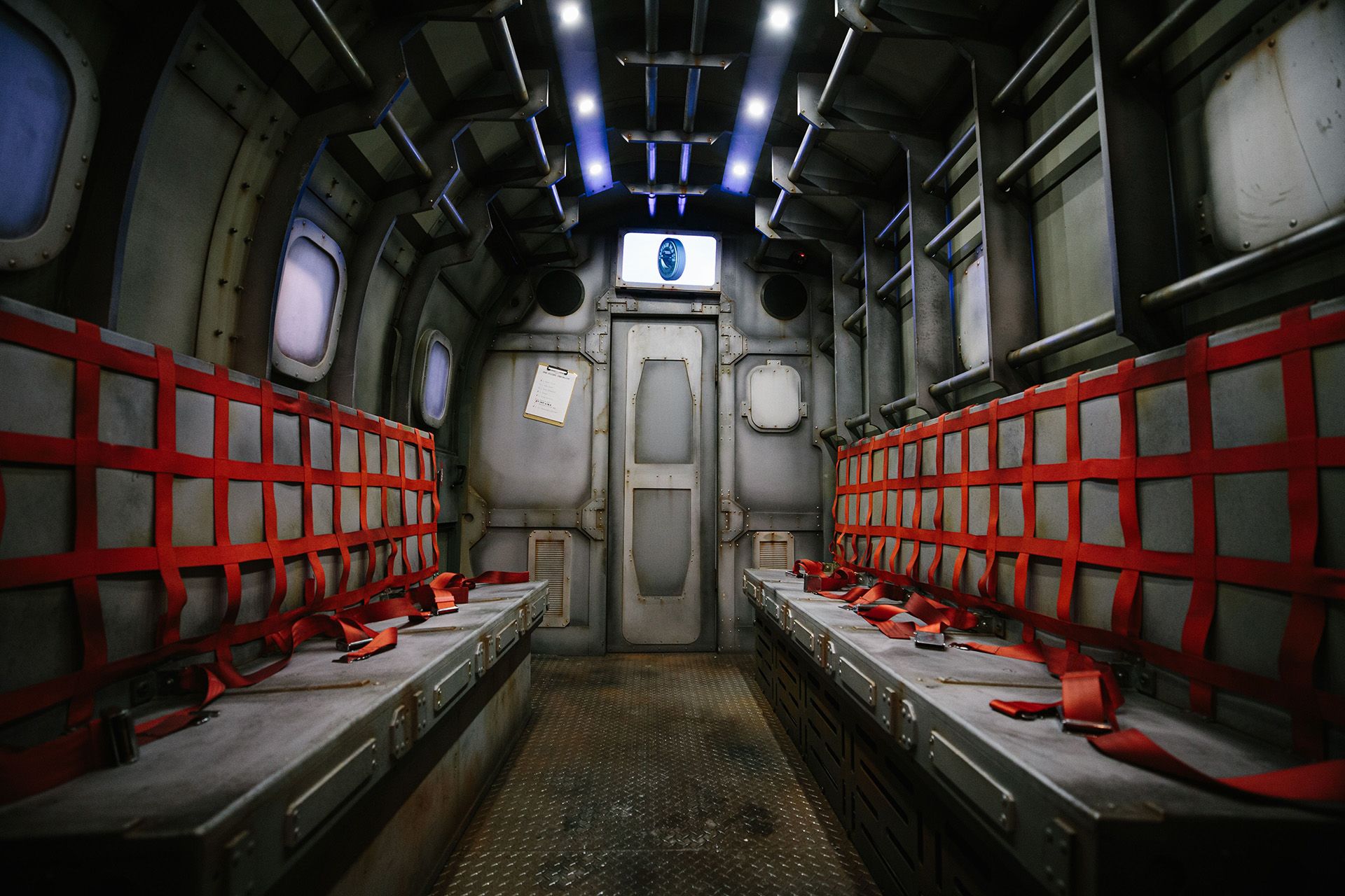 Interior of a futuristic vehicle with metal walls, two rows of seats secured with red straps, and dim lighting.