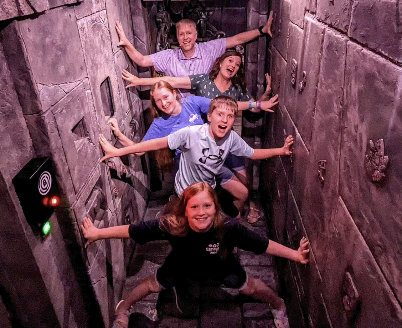 A group of five people poses playfully in a narrow, dimly lit space with textured stone walls, hands outstretched.