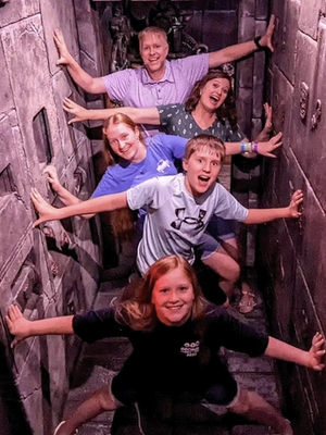 A group of five people poses playfully in a narrow, dimly lit space with textured stone walls, hands outstretched.
