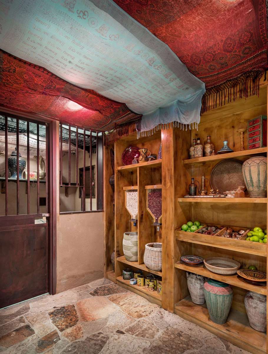 Cozy interior space with wooden shelves filled with pots, baskets, and green apples, under a colorful patterned fabric ceiling.