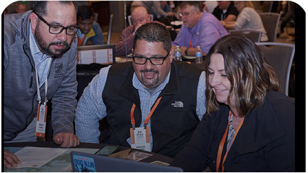 Three individuals are engaged in a discussion over a laptop during a conference. They appear focused and collaborative.