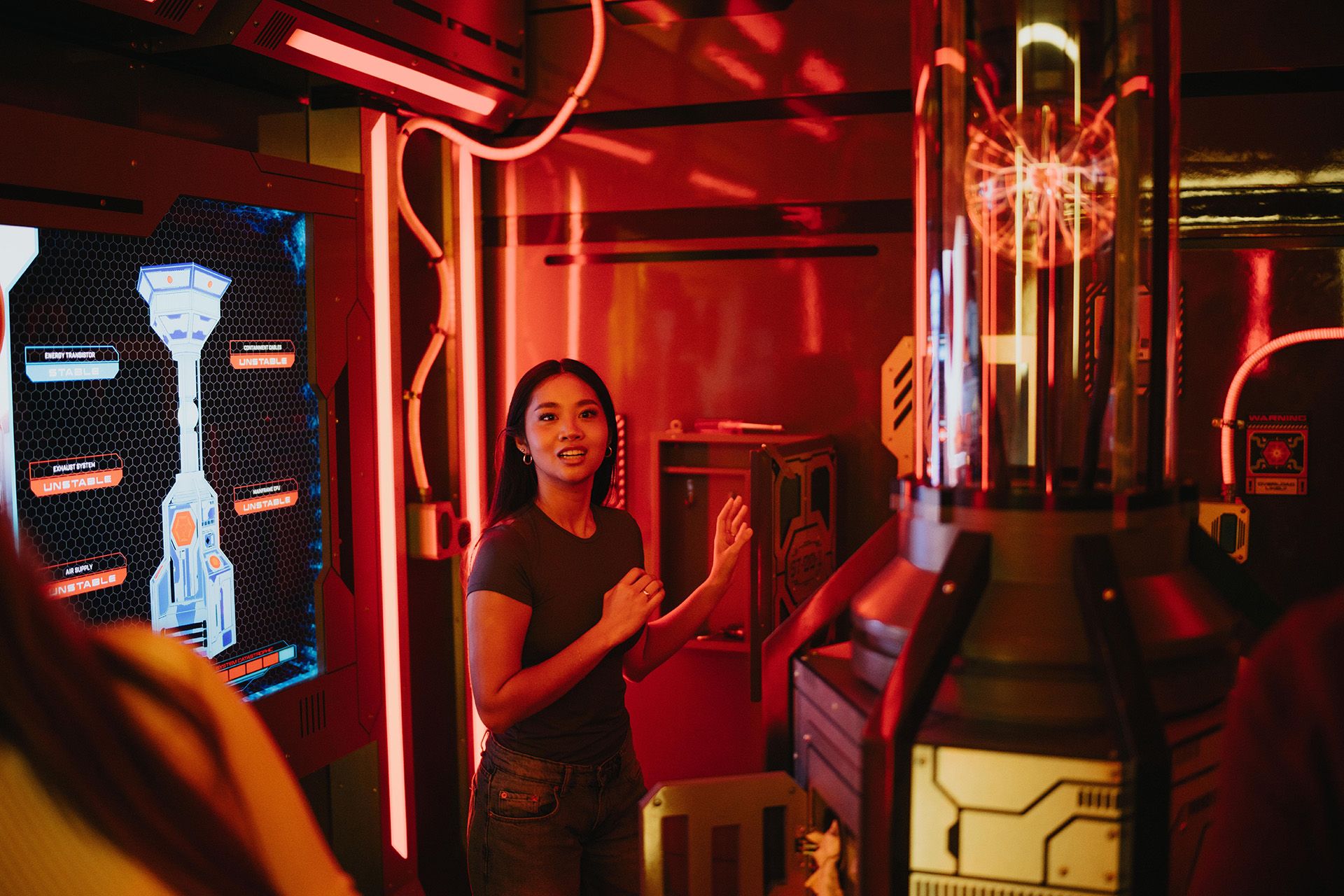 A young woman in a gray shirt gestures animatedly in a futuristic room with red lighting and technology displays.