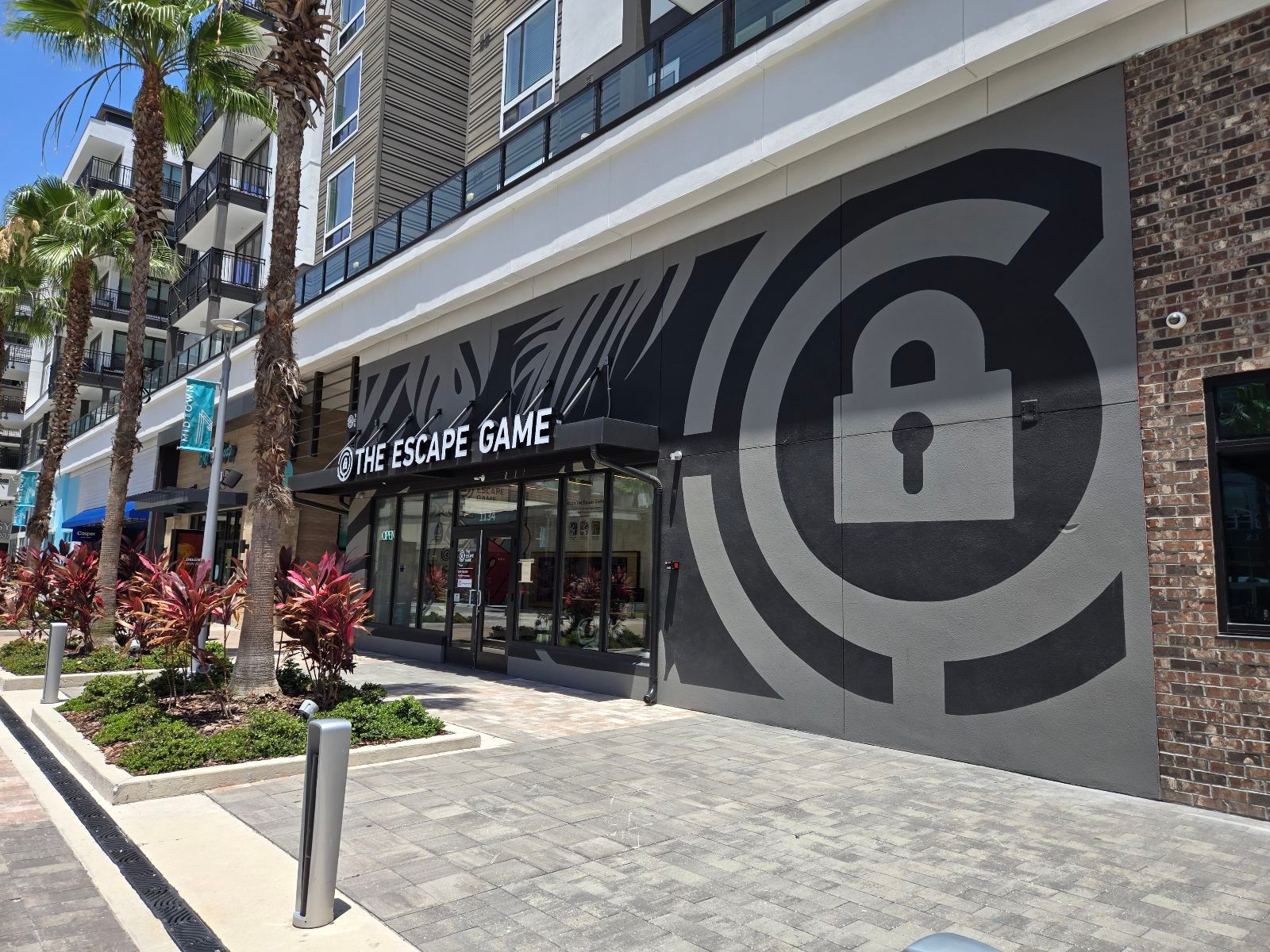 Exterior of "The Escape Game" with a dark wall featuring a lock graphic. Palm trees and colorful plants line the walkway.