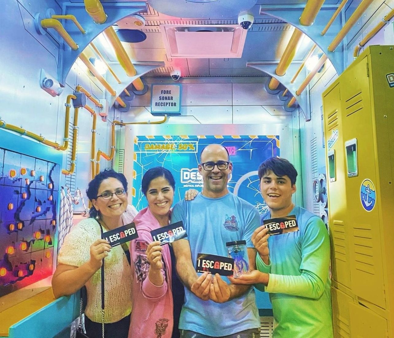 Four people smile and hold "I ESCAPED" signs in a colorful escape room setting with pipes and lockers in the background.