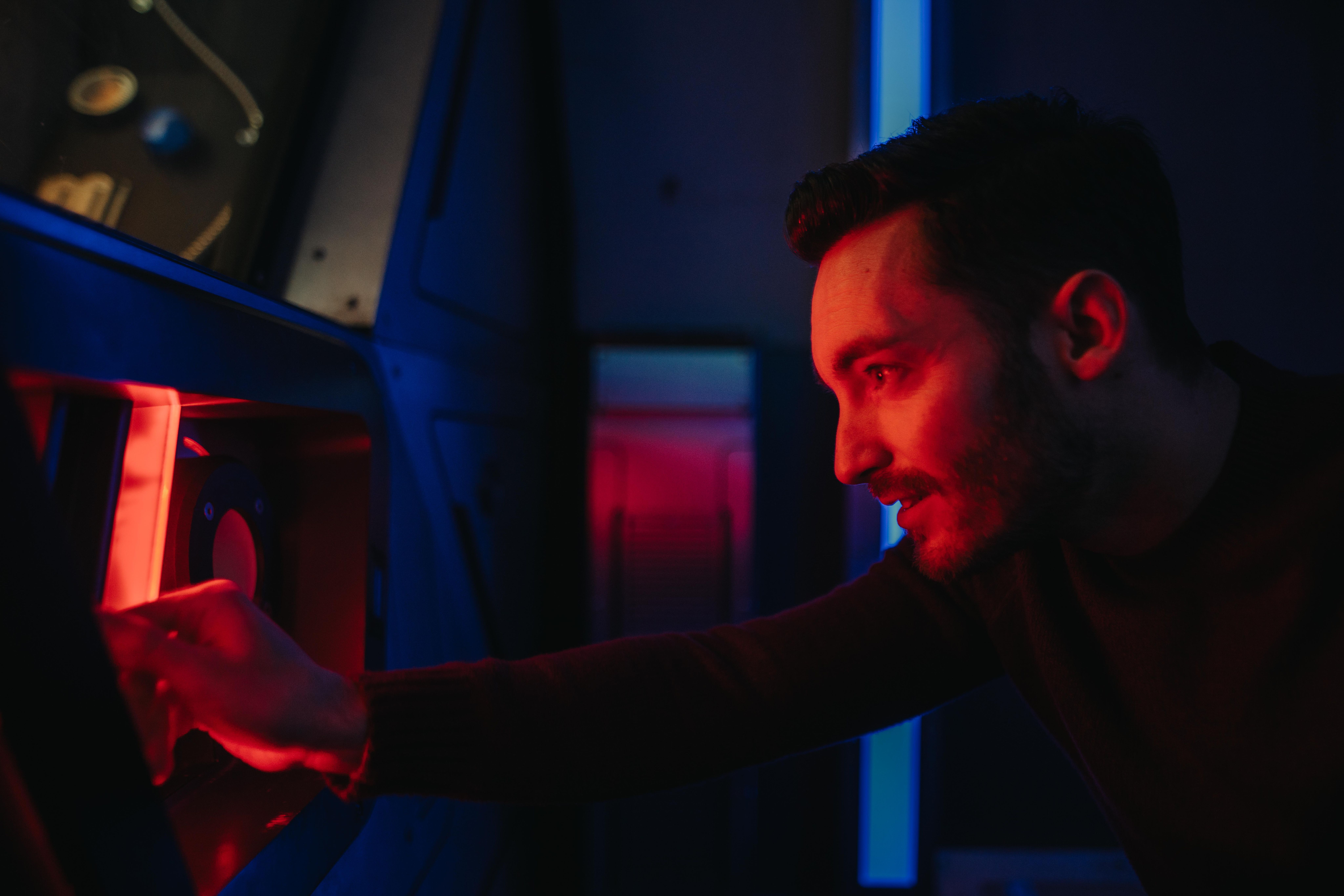 A man leans toward a console with red lighting, focused on an interactive device, in a dimly lit room.